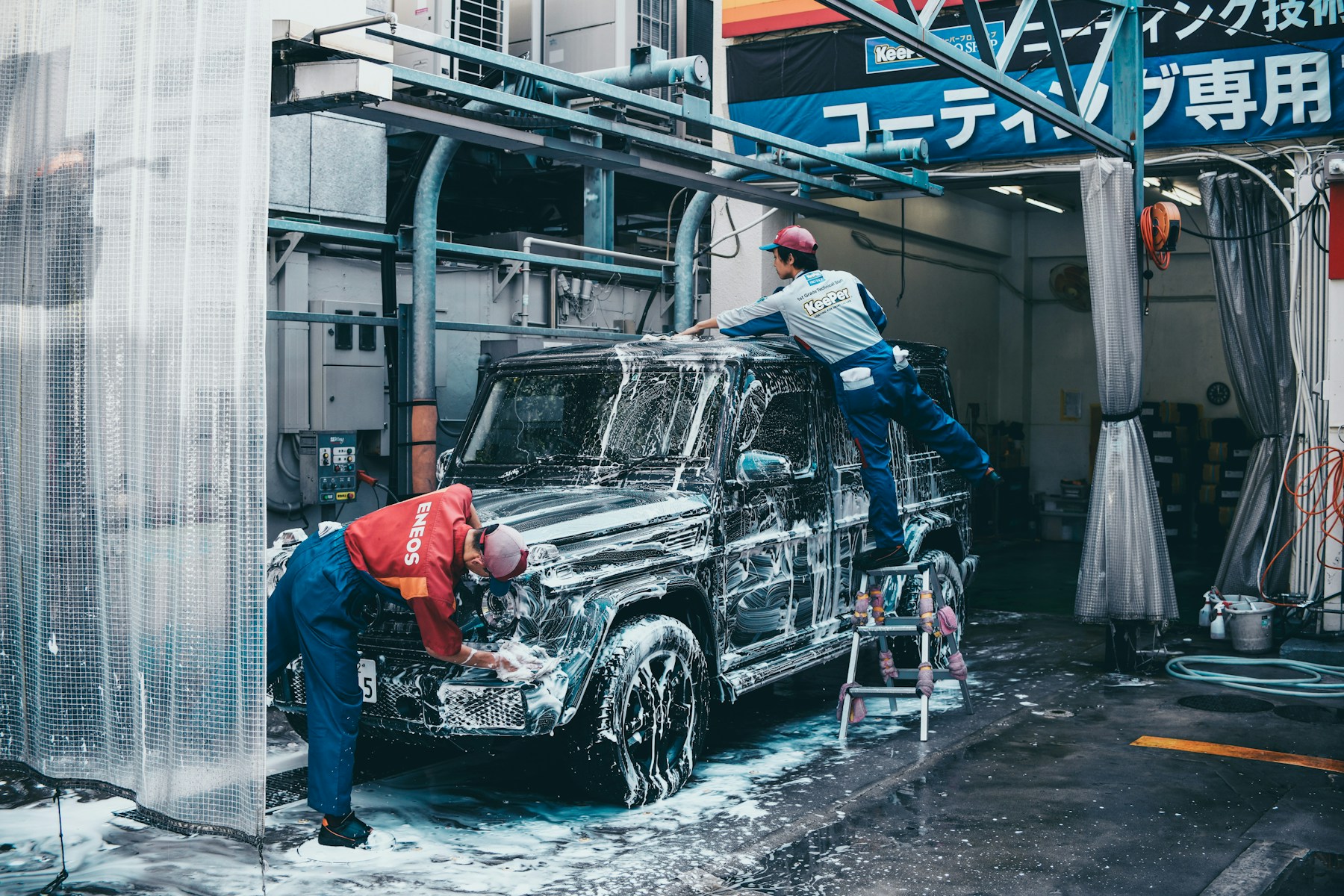 Dua teknisi mencuci SUV hitam penuh busa di bay car wash profesional