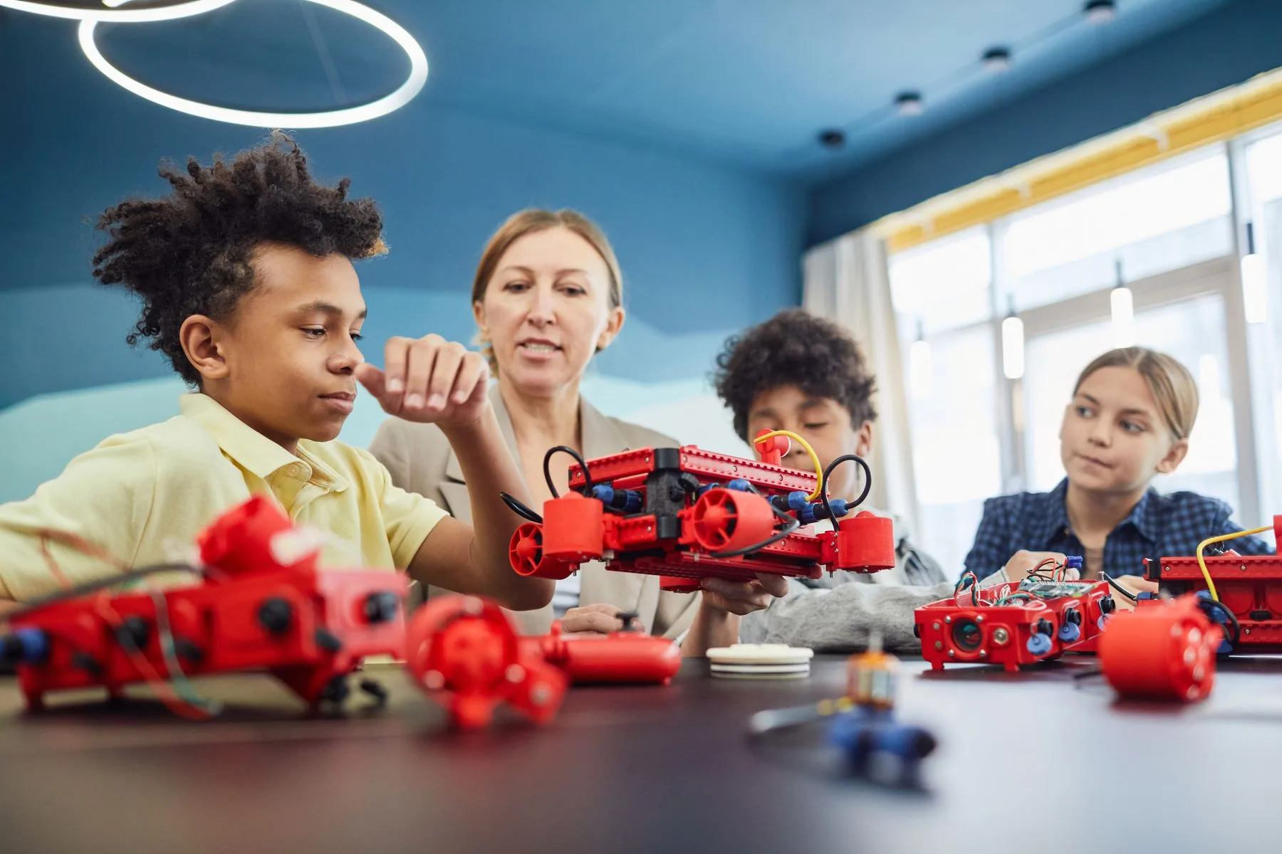 Seorang pengajar mendampingi beberapa anak mengamati rangkaian robot di meja