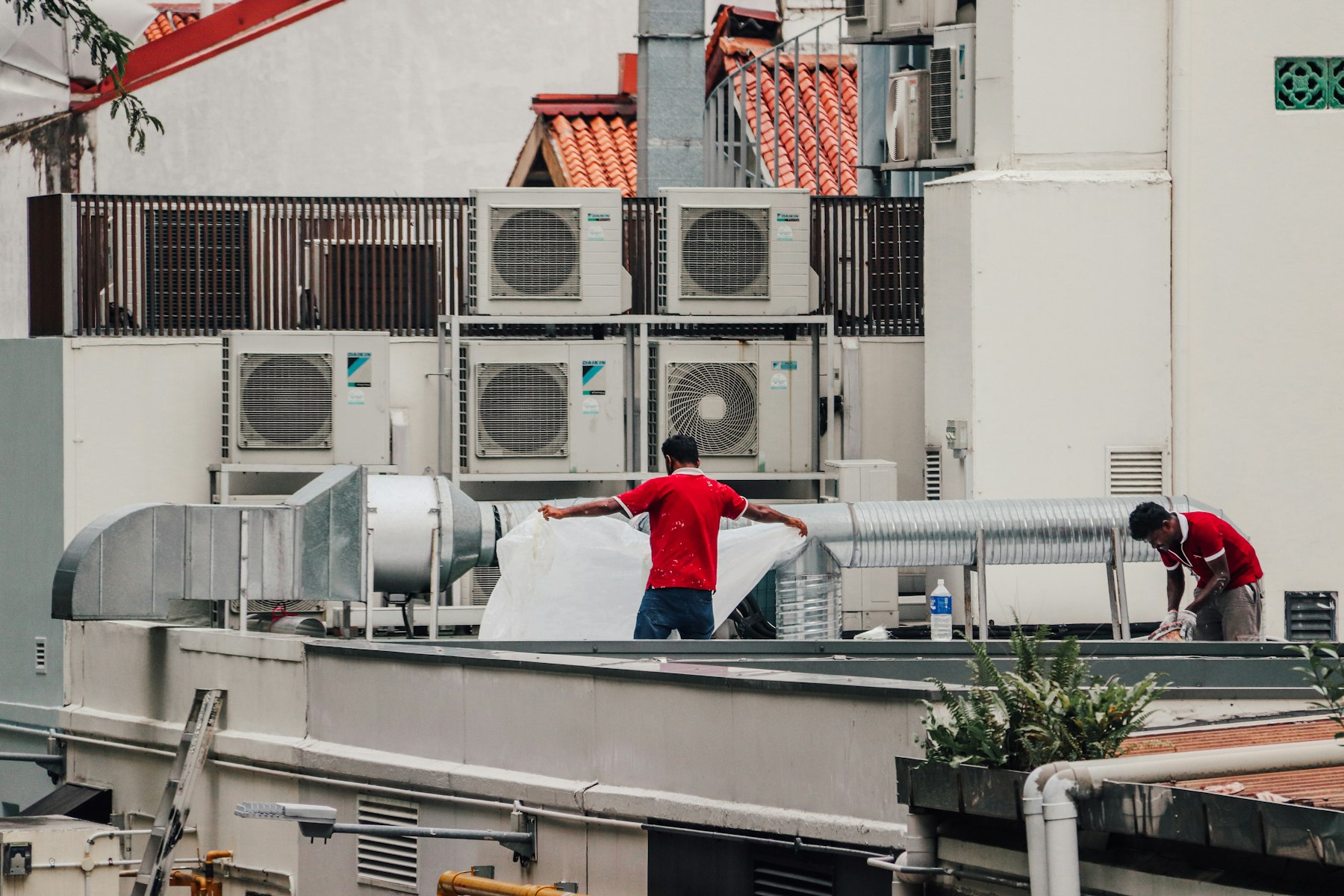 Teknisi HVAC Asia bekerja di rooftop dengan unit AC dan ducting bangunan tropis